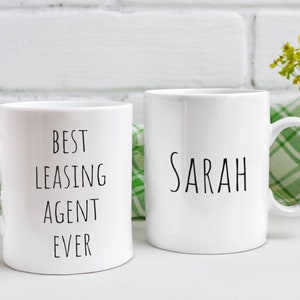 May include: Two white ceramic mugs with black text. One mug says "Best Leasing Agent Ever" and the other mug says "Sarah".