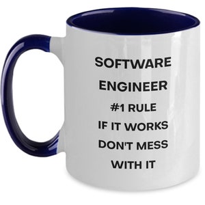 Pu&ograve; includere: Una tazza da caff&egrave; in ceramica bianca con bordo e manico blu. La tazza ha un testo nero che recita "Software Engineer #1 Rule If It Works Don't Mess With It."