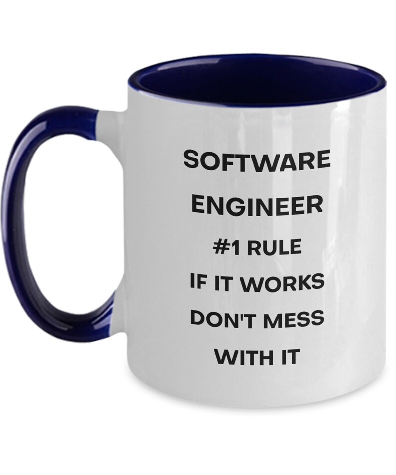 Pu&ograve; includere: Una tazza da caff&egrave; in ceramica bianca con bordo e manico blu. La tazza ha un testo nero che recita "Software Engineer #1 Rule If It Works Don't Mess With It."