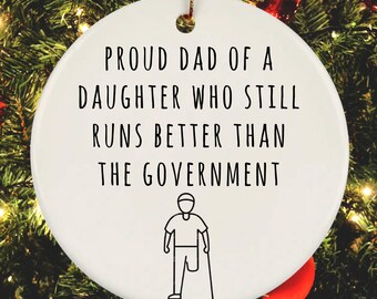 May include: White round Christmas ornament with black text that reads "Proud Dad of a Daughter Who Still Runs Better Than The Government". A stick figure illustration of a person with a prosthetic leg and crutches is below the text.