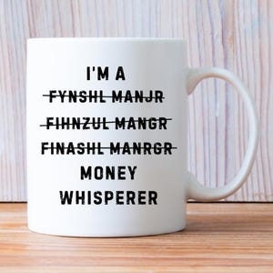 May include: White ceramic mug with the text "I'M A MONEY WHISPERER" in black, with crossed-out words above. The mug has a curved handle and is likely used for beverages. The mug is a humorous gift.