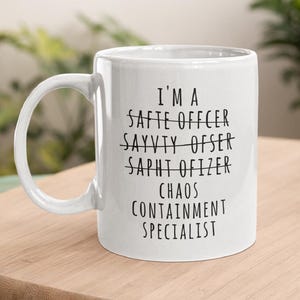 May include: White ceramic mug with a large handle. The mug has the text "I'M A SAFETY OFFICER, CHAOS CONTAINMENT SPECIALIST" printed in black. The words "SAFETY OFFICER" are crossed out and replaced with the correct text.