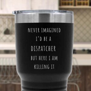 May include: A black insulated tumbler with a silver rim and lid. The tumbler features white text that reads, "NEVER IMAGINED I'D BE A DISPATCHER BUT HERE I AM KILLING IT."