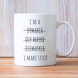 May include: White ceramic mug with a curved handle. The mug features the text "I'M A DYMAKER, DIY MAYKR, DIEMAYKER, I MAKE STUFF" in black, with the first three words crossed out. The mug is on a light brown wooden surface.