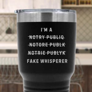 May include: Black stainless steel tumbler with a silver lid. The tumbler has the text "I'M A FAKE WHISPERER" in white, with the words "NOTRY PUBLIQ", "NOTORE PUBLK", and "NOTRIE PUBLYK" crossed out above it.