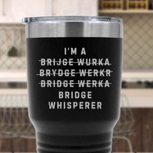 May include: Black insulated tumbler with a silver rim and lid. The tumbler features the text "I'M A BRIDGE WHISPERER" in white, with crossed-out variations of the word "bridge" above it. The background is blurred.