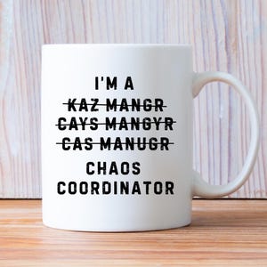 Może przedstawiać: Biały ceramiczny kubek z czarnym napisem "I'M A CHAOS COORDINATOR", z przekreślonymi słowami powyżej. Kubek ma zakrzywiony uchwyt i jest ustawiony na drewnianym tle. Humorystyczny pomysł na prezent.