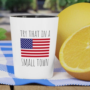 May include: White ceramic mug with a black rim. The mug has a black and red American flag design and the text "TRY THAT IN A SMALL TOWN".