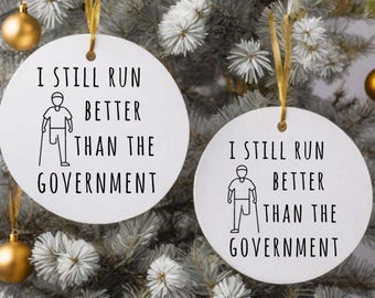 May include: Two white, round Christmas ornaments with gold ribbon hangers. Each ornament has the text "I STILL RUN BETTER THAN THE GOVERNMENT" in black, along with a stick figure illustration.