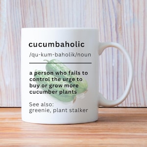 May include: A white ceramic mug featuring the black printed text 'cucumbaholic /qu-kum-baholik/noun' followed by a definition and the phrase 'See also: greenie, plant stalker' centered above a graphic of two green cucumbers.