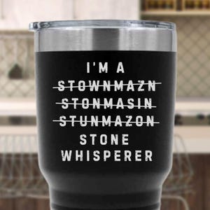 May include: Black stainless steel tumbler with a silver rim and lid. The tumbler features white text that reads "I'M A STOWNMAZN STONMASIN STUNMAZON STONE WHISPERER". The tumbler is designed to keep beverages hot or cold.