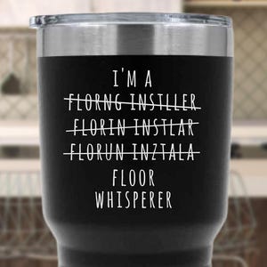 Peut inclure: Un gobelet isotherme noir avec un bord et un couvercle argentés. Le gobelet porte l'inscription blanche "I'M A FLOOR WHISPERER", avec des variantes barrées du mot "installer" au-dessus. Pour boissons chaudes ou froides.