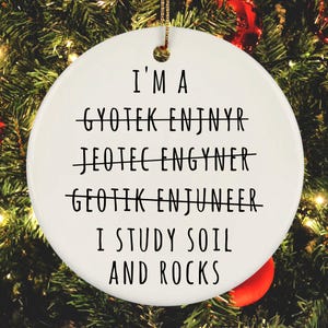 May include: White ceramic Christmas ornament with black text that reads "I'm a Geotek Enjynr, Jeotec Engyner, Geotik Enjuneer, I study soil and rocks".