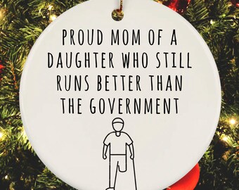 May include: White round Christmas ornament with black text that reads "Proud Mom of a Daughter Who Still Runs Better Than The Government". A black and white illustration of a person with a prosthetic leg and a cane is below the text.