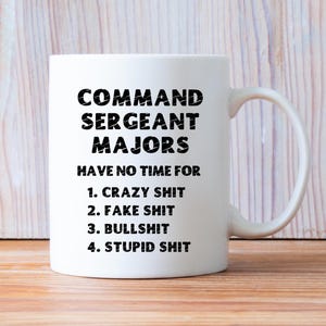 以下が含まれることがあります： 「COMMAND SERGEANT MAJORS HAVE NO TIME FOR 1. CRAZY SHIT 2. FAKE SHIT 3. BULLSHIT 4. STUPID SHIT」と黒い太字で書かれた白いセラミックマグ。 マグカップには曲がったハンドルがあり、明るい木製の背景に置かれています。