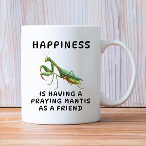 May include: White ceramic mug with a handle. The mug features the word "HAPPINESS" in black, above an illustration of a green praying mantis. Below the insect, the text reads "IS HAVING A PRAYING MANTIS AS A FRIEND."