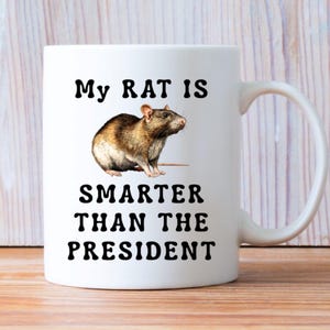 Op de afbeelding: Witte keramische mok met een bruine en bruine rattenillustratie. De mok heeft de tekst "My RAT IS SMARTER THAN THE PRESIDENT" in zwarte, vette letters. De mok heeft een gebogen handvat.