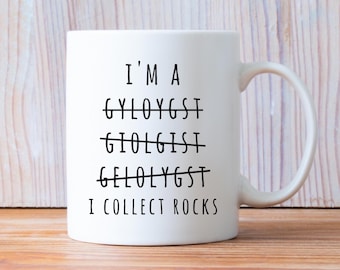 May include: White ceramic mug with the text "I'M A GYLOYGST GIOLGIST GELOLYGST I COLLECT ROCKS" in black. The words "GYLOYGST", "GIOLGIST", and "GELOLYGST" are crossed out. The mug has a curved handle.