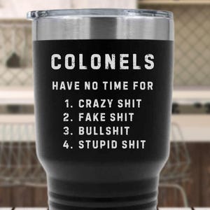 May include: Black stainless steel tumbler with a silver rim and lid. The tumbler has the text "COLONELS HAVE NO TIME FOR 1. CRAZY SHIT 2. FAKE SHIT 3. BULLSHIT 4. STUPID SHIT" in white lettering. The tumbler is on a wooden surface.