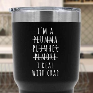 May include: A black stainless steel travel mug with a silver rim and lid. The mug features white text: "I'M A PLUMMA PLUMHER PLMORE I DEAL WITH CRAP". The text is a humorous play on words related to plumbing.