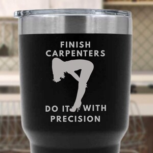 May include: Black stainless steel tumbler with a silver rim. The tumbler features white text that reads "FINISH CARPENTERS DO IT WITH PRECISION" with a silhouette graphic of a person bending over.