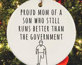 May include: White round Christmas ornament with black text that reads "Proud Mom of a Son Who Still Runs Better Than The Government". A black and white illustration of a person with a prosthetic leg and crutches is below the text.