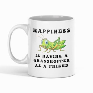 May include: White ceramic mug with a green grasshopper illustration and the text "HAPPINESS IS HAVING A GRASSHOPPER AS A FRIEND" in black. The grasshopper is smiling with blue eyes.
