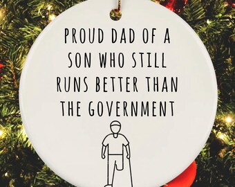 May include: White ceramic Christmas ornament with black text that reads "Proud Dad of a Son Who Still Runs Better Than The Government". A black and white illustration of a person with a prosthetic leg and a cane is below the text.