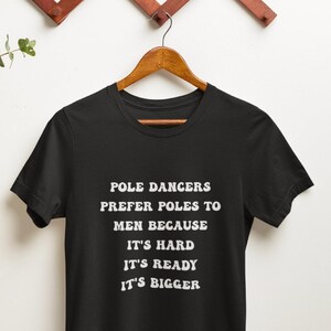 May include: A black short-sleeved t-shirt featuring white text that reads: POLE DANCERS PREFER POLES TO MEN BECAUSE IT'S HARD IT'S READY IT'S BIGGER. The shirt is displayed on a wooden hanger, showcasing a crew neckline and a standard fit design.
