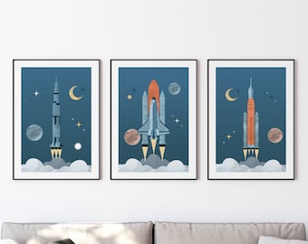 May include: Three framed prints with a blue background and white stars. Each print features a different rocket ship in flight. The first print shows a white rocket with an orange tip. The second print shows a white rocket with a blue and orange tip. The third print shows a white rocket with a red and orange tip.