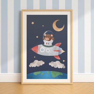 May include: A framed art print depicting a brown bear astronaut in a rocket ship against a dark blue space backdrop. The rocket is grey with red fins, and the scene includes a crescent moon, stars, clouds, and a blue and green planet.