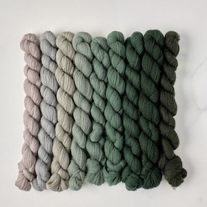 May include: A set of ten skeins of yarn in shades of green, gray, and beige. The yarn is arranged in a gradient from light to dark.