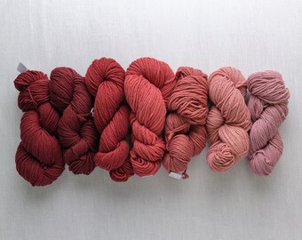 Puede incluir: Siete madejas de hilo en un degradado de tonos rojos, desde el burdeos oscuro hasta el rosa claro. El hilo está bien enrollado y dispuesto horizontalmente sobre un fondo neutro y texturizado. Ideal para proyectos de punto y ganchillo.