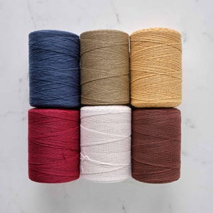 May include: Seven spools of colorful macrame cord in shades of blue, tan, yellow, red, white, and brown. The spools are arranged in two rows, with the colors varying. The cord is tightly wound and ready for crafting projects.