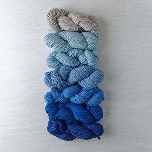 May include: A stack of yarn in shades of blue and gray. The yarn is arranged from lightest to darkest, with the gray yarn at the top and the darkest blue yarn at the bottom.
