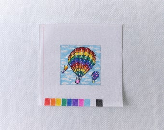 May include: A white canvas with a colorful hot air balloon design. The balloon is rainbow colored with a blue sky and white clouds in the background. There are two smaller hot air balloons in the background, one yellow and one purple.