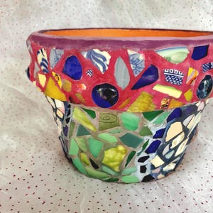 May include: A round flower pot decorated with a mosaic of colorful ceramic and glass shards. The rim features a vibrant pink grout with blue, yellow, and white pieces, while the body displays a mix of green, blue, and white glass fragments.