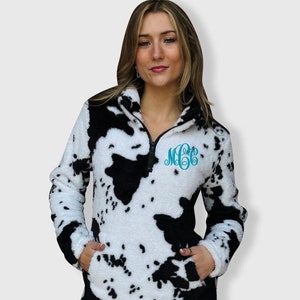 May include: A white and black cow print fleece pullover with a quarter zip closure and a monogrammed "MGE" on the chest.