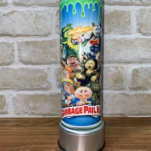 Puede incluir: Un vaso de acero inoxidable con un diseño azul y verde con personajes de Garbage Pail Kids. El diseño incluye una baba verde que gotea por el lado del vaso. El texto "Garbage Pail Kids" está impreso en la parte inferior del vaso.