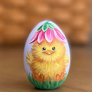 May include: A hand-painted Easter egg with a yellow chick wearing a pink flower on its head. The chick is standing in green grass.