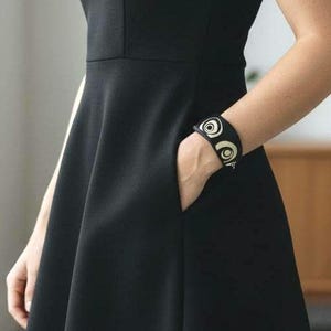 Op de afbeelding: Een zwarte mouwloze jurk met een getailleerd lijfje en een uitlopende rok. De jurk heeft een ronde halslijn en een subtiel naaddetail. Een zwarte armband met een cirkelvormig ontwerp is zichtbaar op de pols.