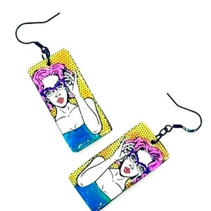 May include: A pair of rectangular earrings with a pop art design. Each earring displays a woman with pink hair and glasses, set against a yellow backdrop with black dots and a blue and white design.