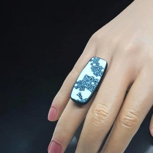 May include: A rectangular ring with a light blue and dark blue patterned design. The ring is set on a black band. The ring is worn on a finger.