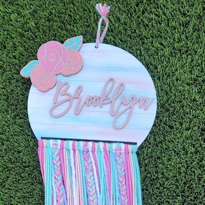 May include: A pink, white, and blue yarn wall hanging with a round wooden sign that says "Brooklyn" with a floral design.