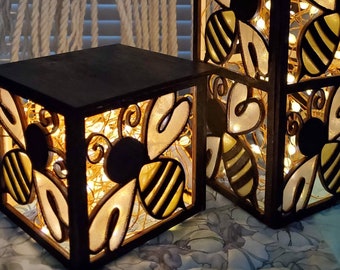 Puede incluir: Dos cajas de luz de madera con temática de abejas con luces de cadena en el interior. La caja más grande es más alta y tiene tres secciones con abejas y corazones recortados. La caja más pequeña es cuadrada y tiene una sección con abejas y corazones recortados.