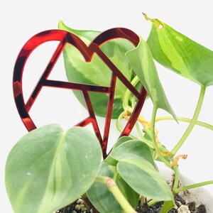 May include: A red heart-shaped plant stake with a geometric design is placed in a pot with a green leafy plant.