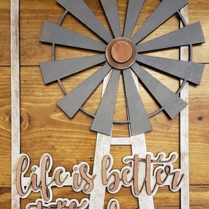 Könnte beinhalten: Ein Holzschild mit einem weiß getünchten Rahmen und einem verwitterten Aussehen. Das Schild zeigt eine Windmühle aus Metall mit einem kupfernen Zentrum und den Worten "life is better at the farm" in kupfernen Lettern.