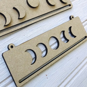 May include: Two wooden moon phase organizers with cutouts of different moon phases. The organizers are made of light brown wood and have a black border.