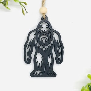 May include: Black wooden ornament shaped like a Bigfoot, a mythical creature with large feet and hairy body. The ornament is hanging from a string with a wooden bead.