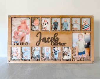 May include: A wooden photo frame with 12 individual picture slots, each with a label for a different month of the year. The frame is decorated with the name "Jacob Carter" and the words "Hello", "One Year", "Two Weeks", "One Month", "Two Months", "Three Months", "Four Months", "Five Months", "Six Months", "Seven Months", "Eight Months", "Nine Months", "Ten Months", "Eleven Months".